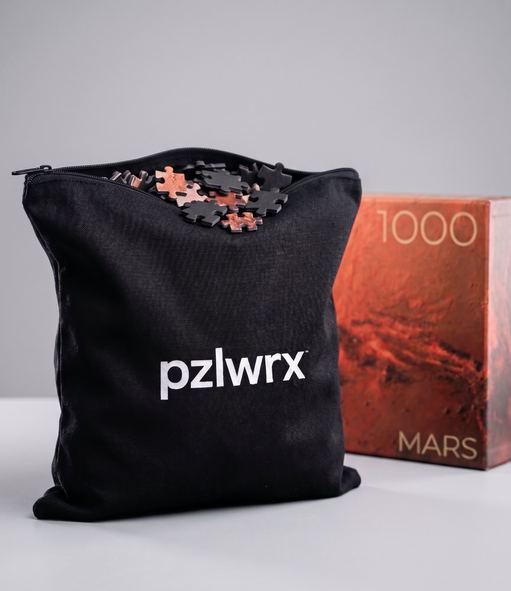 A black canvas pzlwrx zip pouch filled with puzzle pieces beside the rust-red Mars 1000-piece box